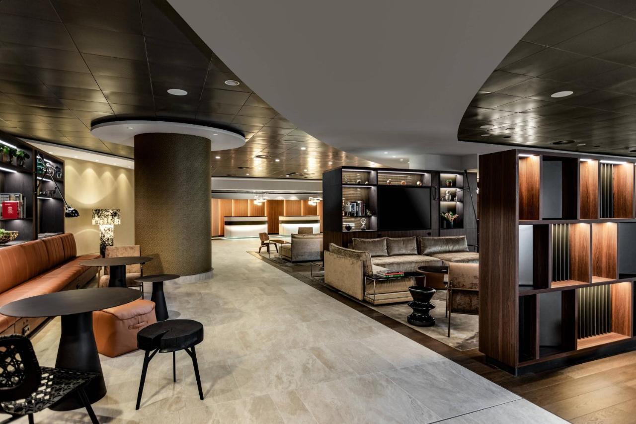 Frankfurt Airport Marriott Hotel Φραγκφούρτη Εξωτερικό φωτογραφία The photo shows a modern and stylish interior space, likely a hotel lobby or lounge. It features curved walls and a sleek ceiling design. There are comfortable seating areas with a mix of sofas and chairs arranged around small tables. A shelving unit