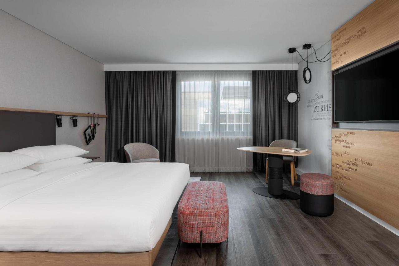 Frankfurt Airport Marriott Hotel Φραγκφούρτη Εξωτερικό φωτογραφία The photo shows a modern hotel room featuring a minimalist design. There's a large bed with white linens, flanked by two bedside tables. In the corner, there are two chairs next to a small round table. A flat-screen TV is mounted on the wall opposite