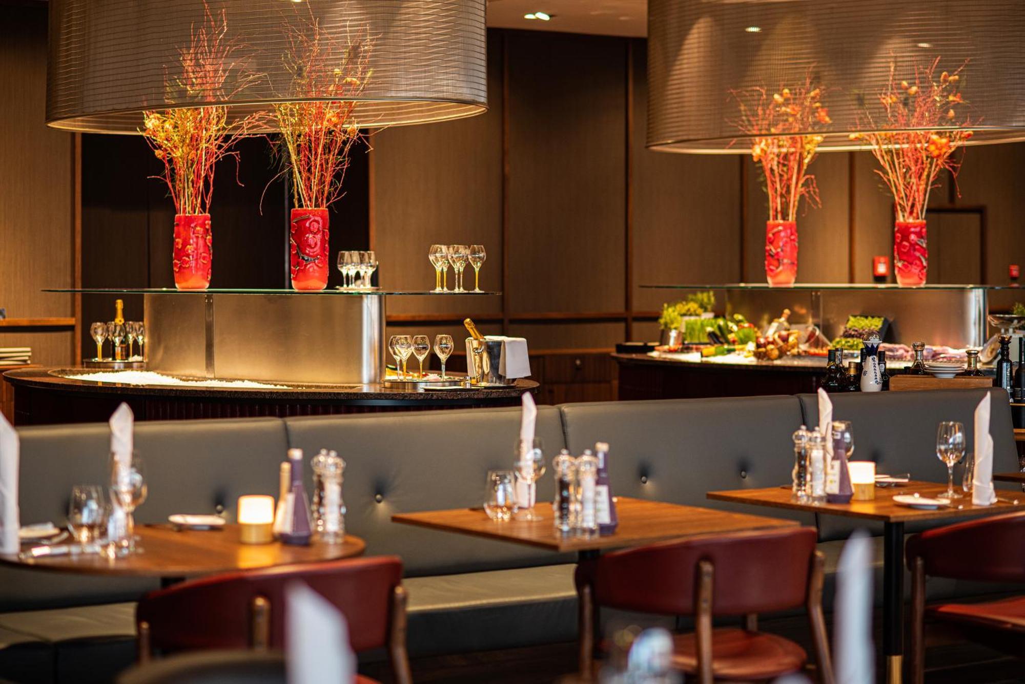 Frankfurt Airport Marriott Hotel Φραγκφούρτη Εξωτερικό φωτογραφία The photo shows an elegant restaurant interior. It features wooden tables with chairs arranged neatly, creating a warm and inviting atmosphere. In the background, there are large decorative vases with colorful floral arrangements, adding a pop of col