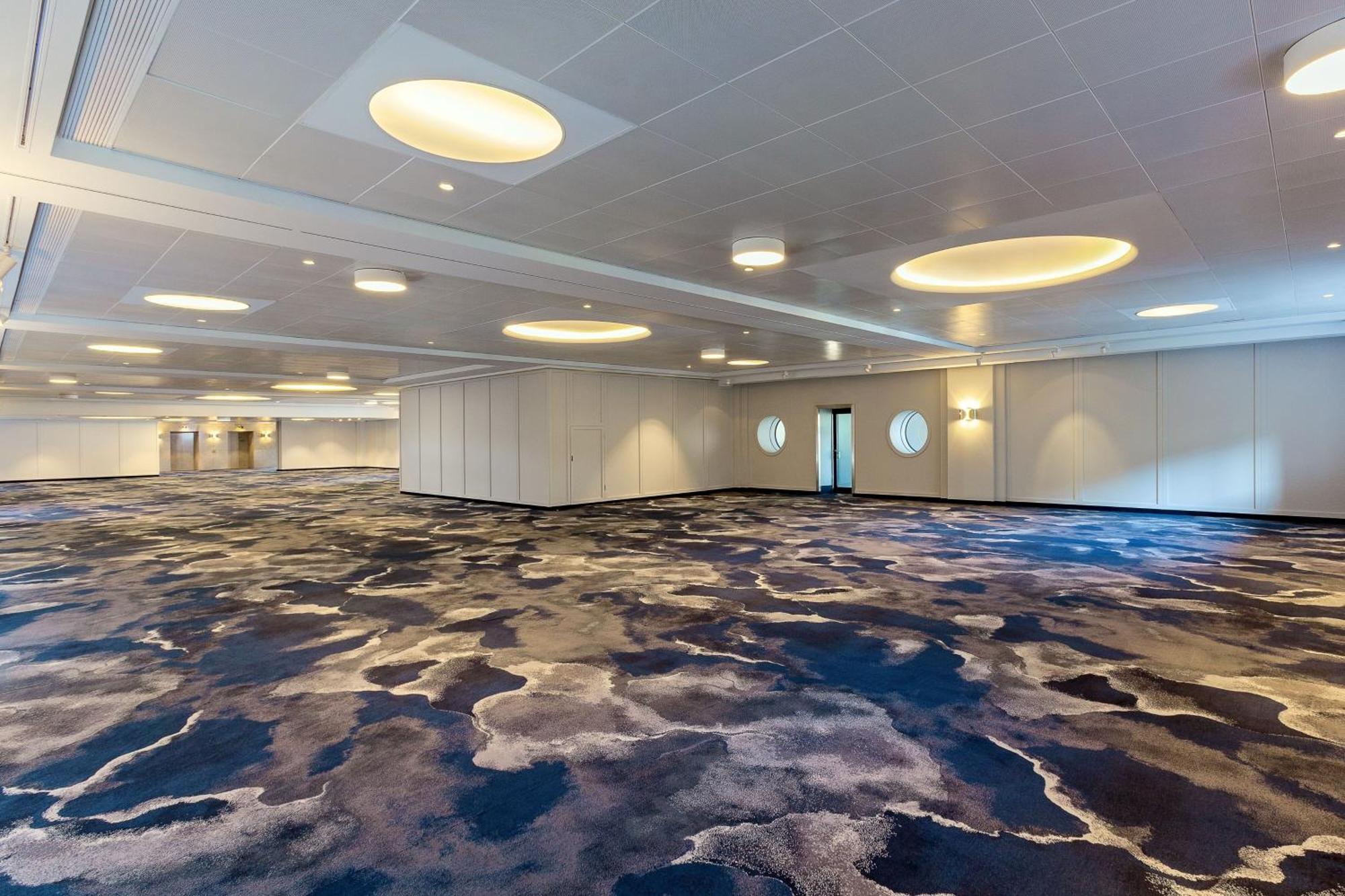 Frankfurt Airport Marriott Hotel Φραγκφούρτη Εξωτερικό φωτογραφία The photo shows a spacious, well-lit interior room or hall. The flooring features a distinctive carpet design with abstract patterns in shades of blue and gray. The ceiling has large, circular light fixtures that provide ambient lighting. There are a