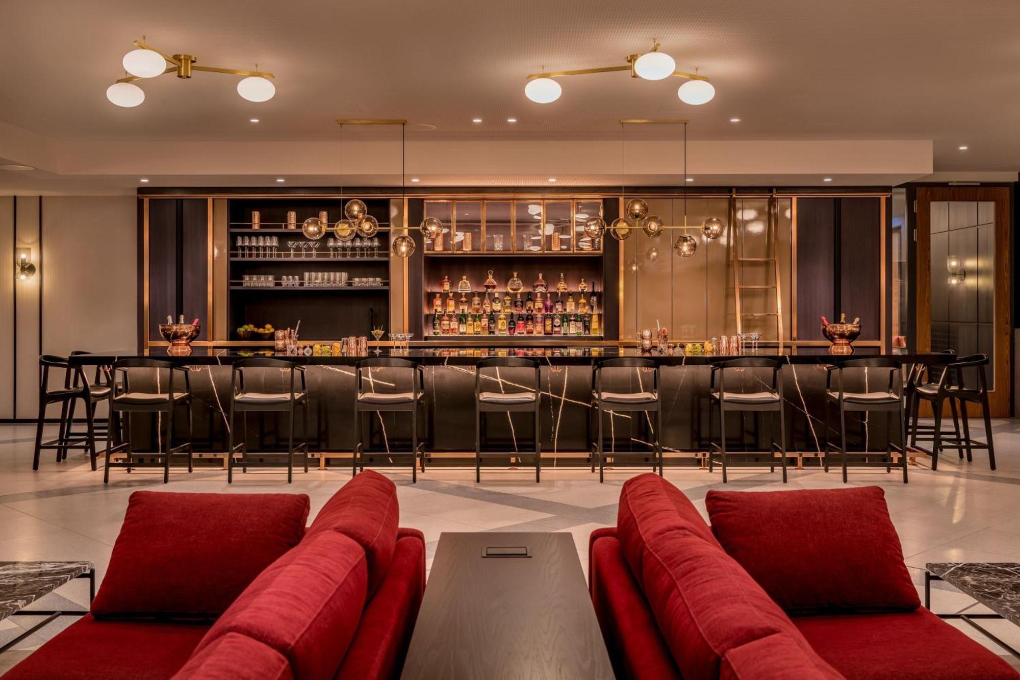 Frankfurt Airport Marriott Hotel Φραγκφούρτη Εξωτερικό φωτογραφία The photo shows a stylish bar area. In the foreground, there are red sofas arranged for seating. Behind the seating area, there is a long bar with a dark countertop. The bar is well-stocked with various bottles displayed on shelves. Elegant lighting 
