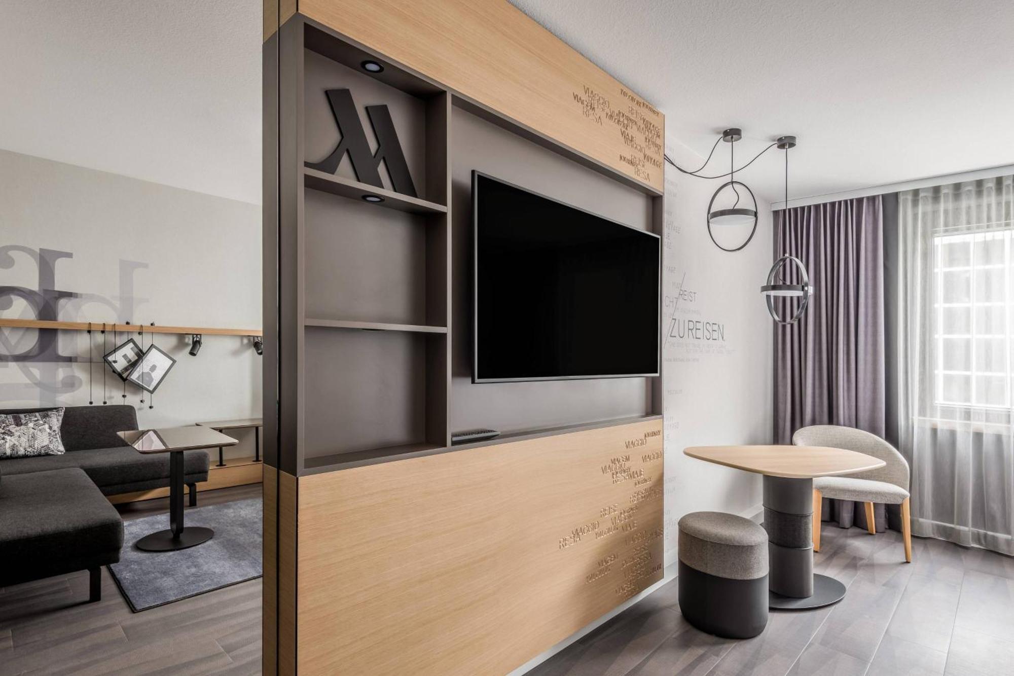 Frankfurt Airport Marriott Hotel Φραγκφούρτη Εξωτερικό φωτογραφία The photo shows a contemporary hotel room or living space. It features a wooden accent wall with built-in shelves, containing decorative items and a television set. There is a small dining table with two upholstered stools nearby. The space is well-l