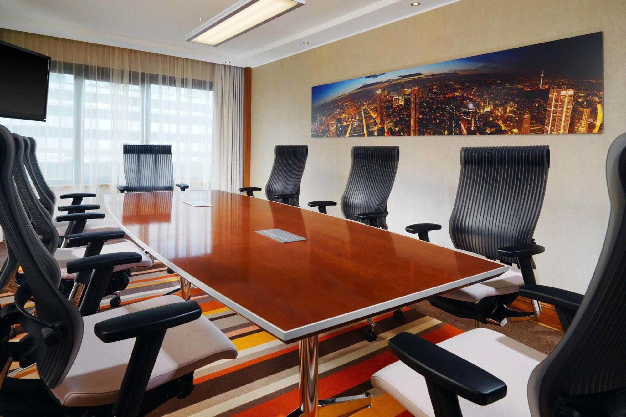 Frankfurt Airport Marriott Hotel Φραγκφούρτη Εξωτερικό φωτογραφία The photo depicts a modern conference room featuring a large wooden table surrounded by ergonomic black office chairs. The room is well-lit with large windows that allow natural light to enter, and there is a panoramic image of a city skyline on the 