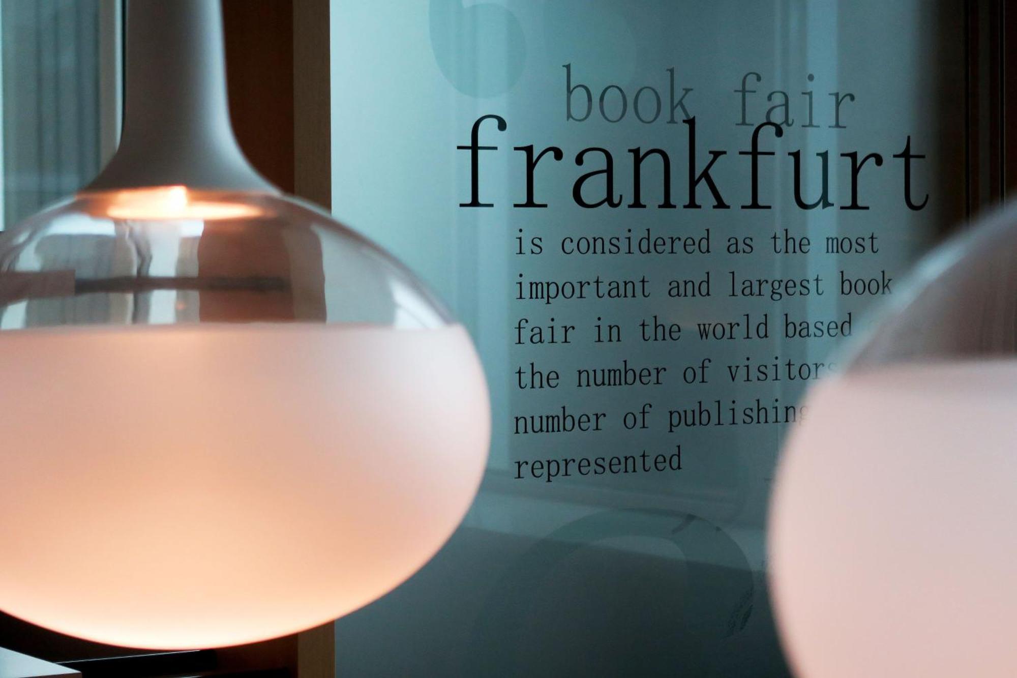 Frankfurt Airport Marriott Hotel Φραγκφούρτη Εξωτερικό φωτογραφία The photo features two modern pendant light fixtures hanging from the ceiling. In the background, there is a piece of glass or wall with text that reads about the Frankfurt Book Fair, stating that it is the most important and largest book fair in the