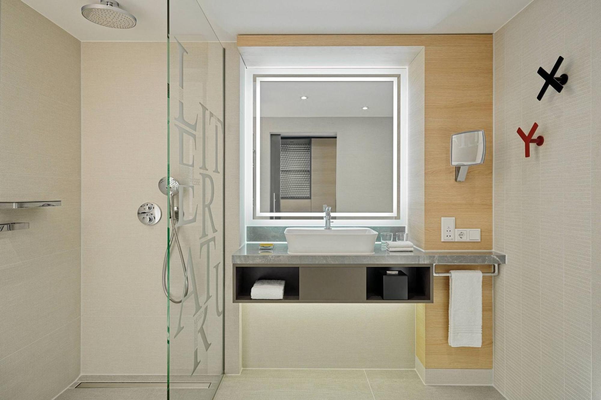 Frankfurt Airport Marriott Hotel Φραγκφούρτη Εξωτερικό φωτογραφία The photo shows a modern bathroom with a sleek design. It features a glass shower enclosure on the left, a stylish sink with a large mirror above it, and a countertop underneath the mirror. The wall behind the sink is made of light-colored wood, enha