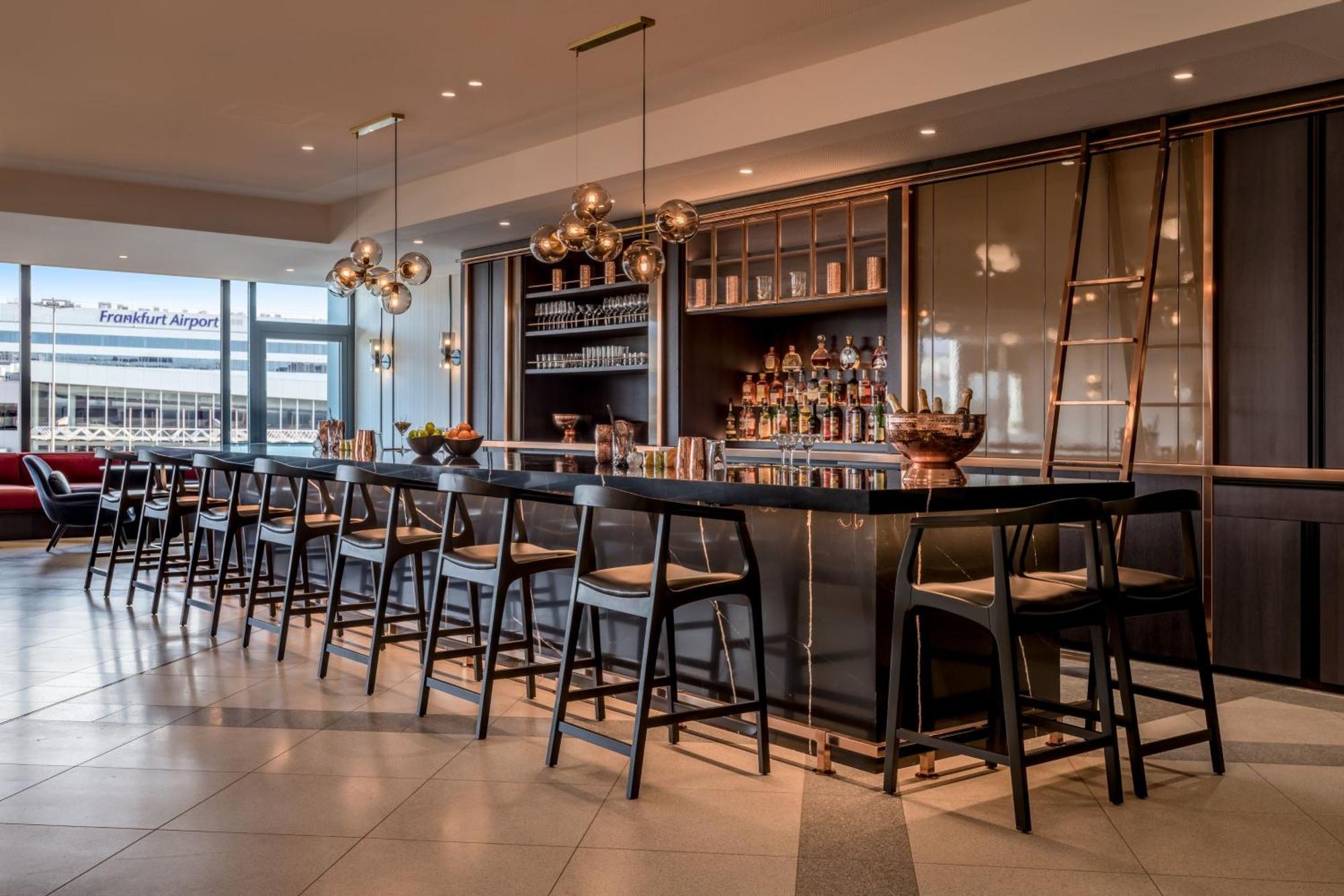Frankfurt Airport Marriott Hotel Φραγκφούρτη Εξωτερικό φωτογραφία The photo shows a modern bar area with a sleek design. There is a long dark bar counter with several high stools placed along it. Above the bar, stylish pendant lights provide warm illumination. Behind the bar, a shelf holds various bottles of liquor