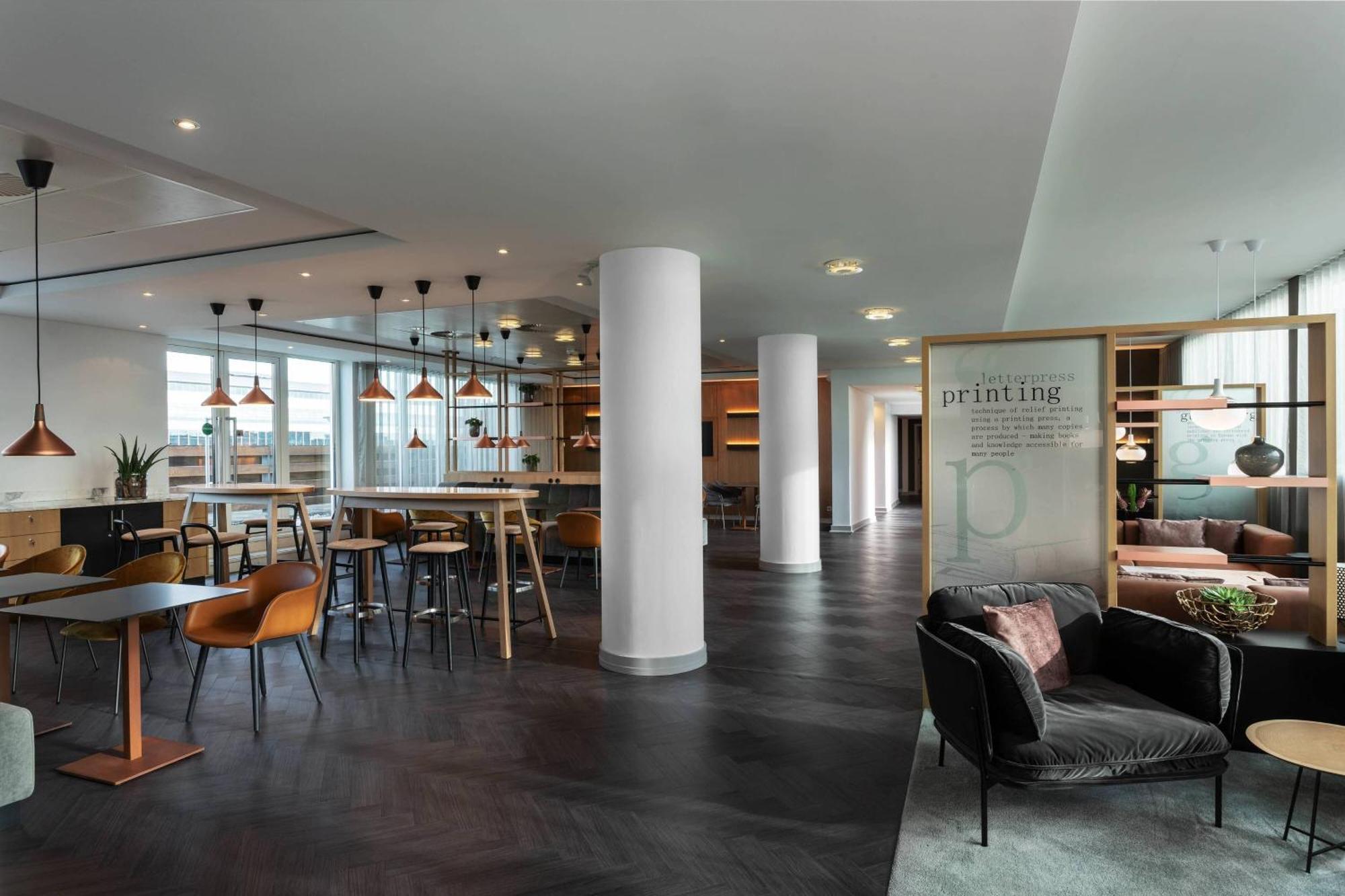Frankfurt Airport Marriott Hotel Φραγκφούρτη Εξωτερικό φωτογραφία The photo shows a modern, stylish interior of a workspace or a lounge area. It features a mix of seating arrangements, including a cozy dark sofa and tables with chairs. The space has an open layout with sleek flooring, large windows allowing natural